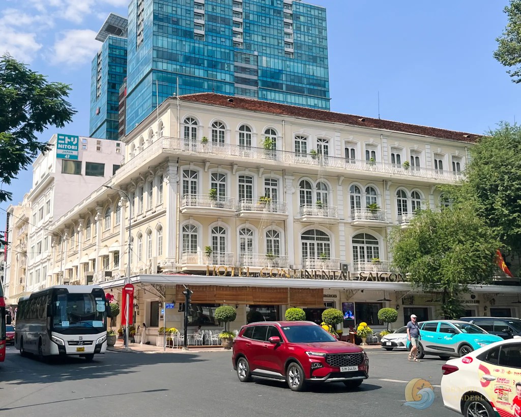 A busy street scene featuring the Hotel Continental Saigon, showcasing its elegant white facade with ornate balconies, surrounded by modern buildings. Various vehicles, including a bus and cars, are seen on the road, with pedestrians nearby and greenery adding a lively atmosphere.