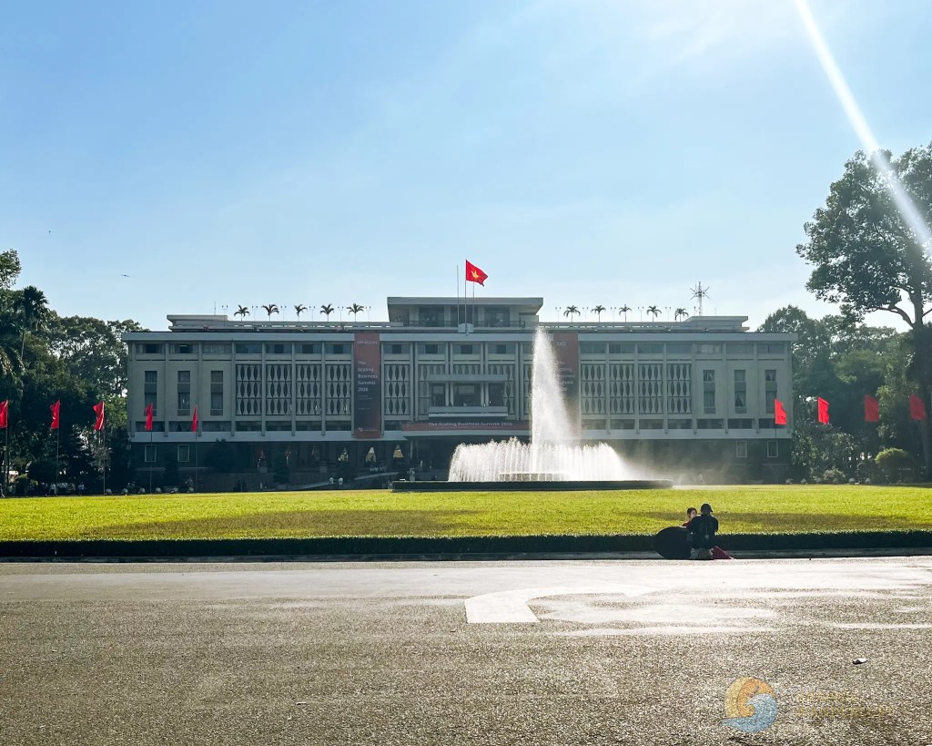 The Reunification Palace in Ho Chi Minh City, Vietnam, featuring a fountain and surrounded by greenery and red flags under a clear blue sky.