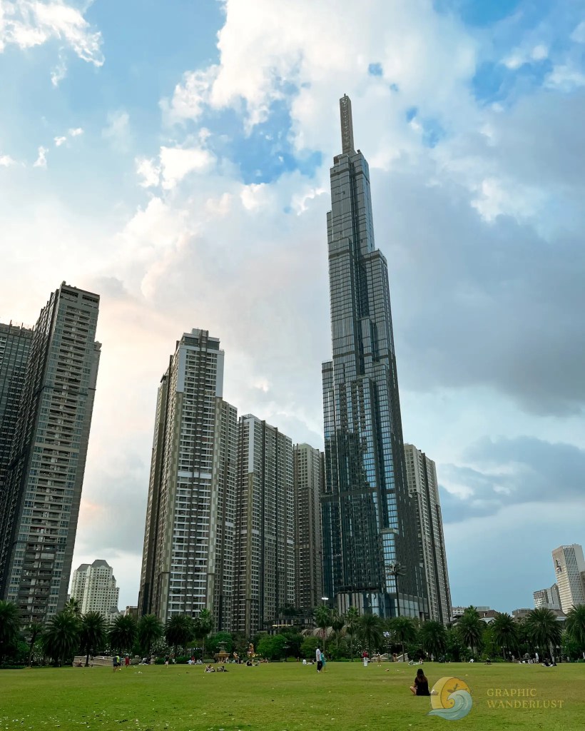 Skyscraper towering over modern buildings and a green park, with people enjoying the open space under a cloudy sky.