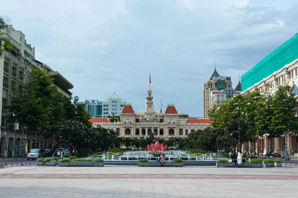 A wide view of a public square featuring a fountain and flowering plants, with a prominent historical building in the background, surrounded by modern skyscrapers and greenery under a partly cloudy sky.