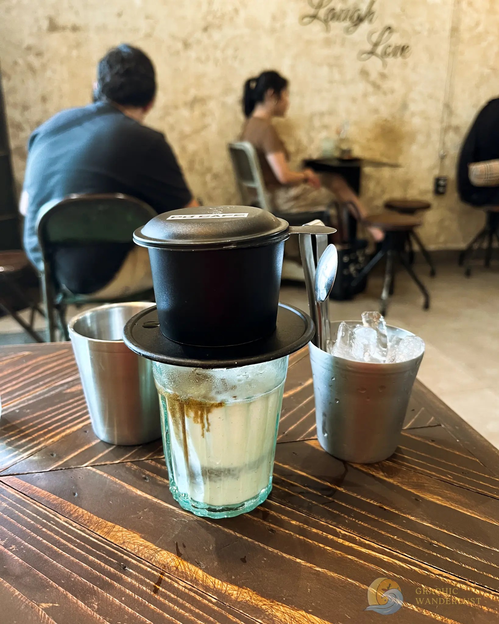 A serving drip-over coffee placed on top of milk-filled clear glass with two metal glasses beside it and dining customers in the background. 
