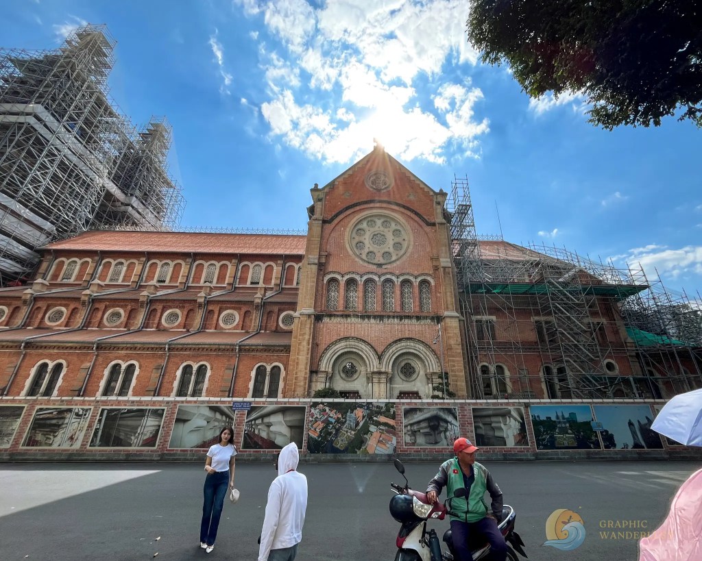 A partially renovated historic church building with scaffolding, surrounded by blue skies and scattered clouds, featuring two people in the foreground and a motorcycle parked nearby.