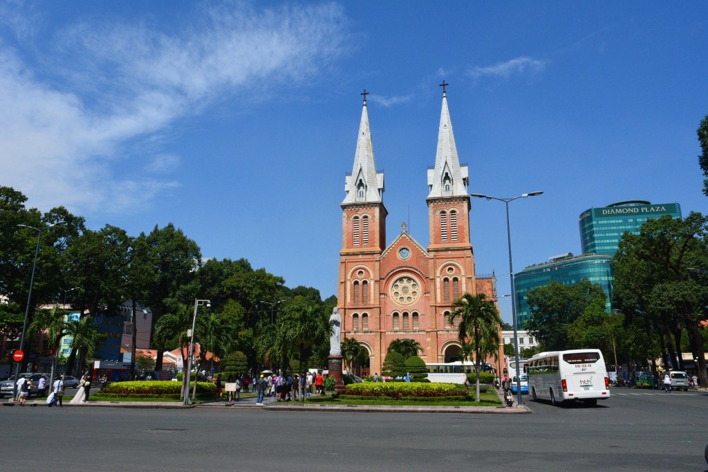 A view of the Saigon Notre-Dame Basilica, featuring its distinctive twin spires, surrounded by greenery and bustling streets, under a clear blue sky.