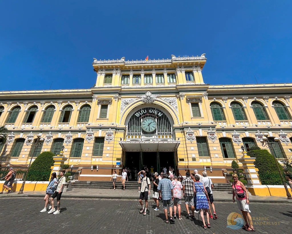 A historic yellow and white postal building with a large clock and arched entrance, surrounded by trees and tourists on a sunny day.