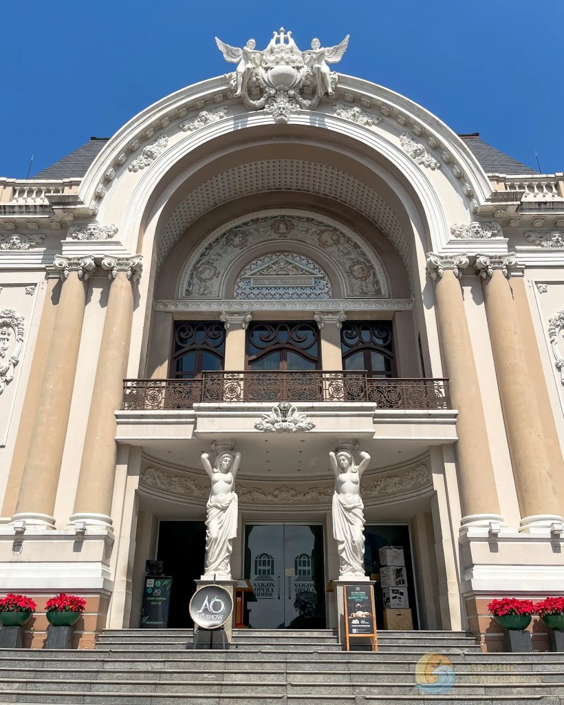 The entrance of a grand building featuring ornate architecture, including two statues of women on either side of the stairs, decorated with vibrant red flowers.