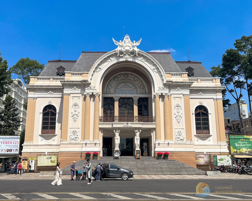 Exterior view of an ornate theater building with a large arched entrance, decorative statues, and a clear blue sky.