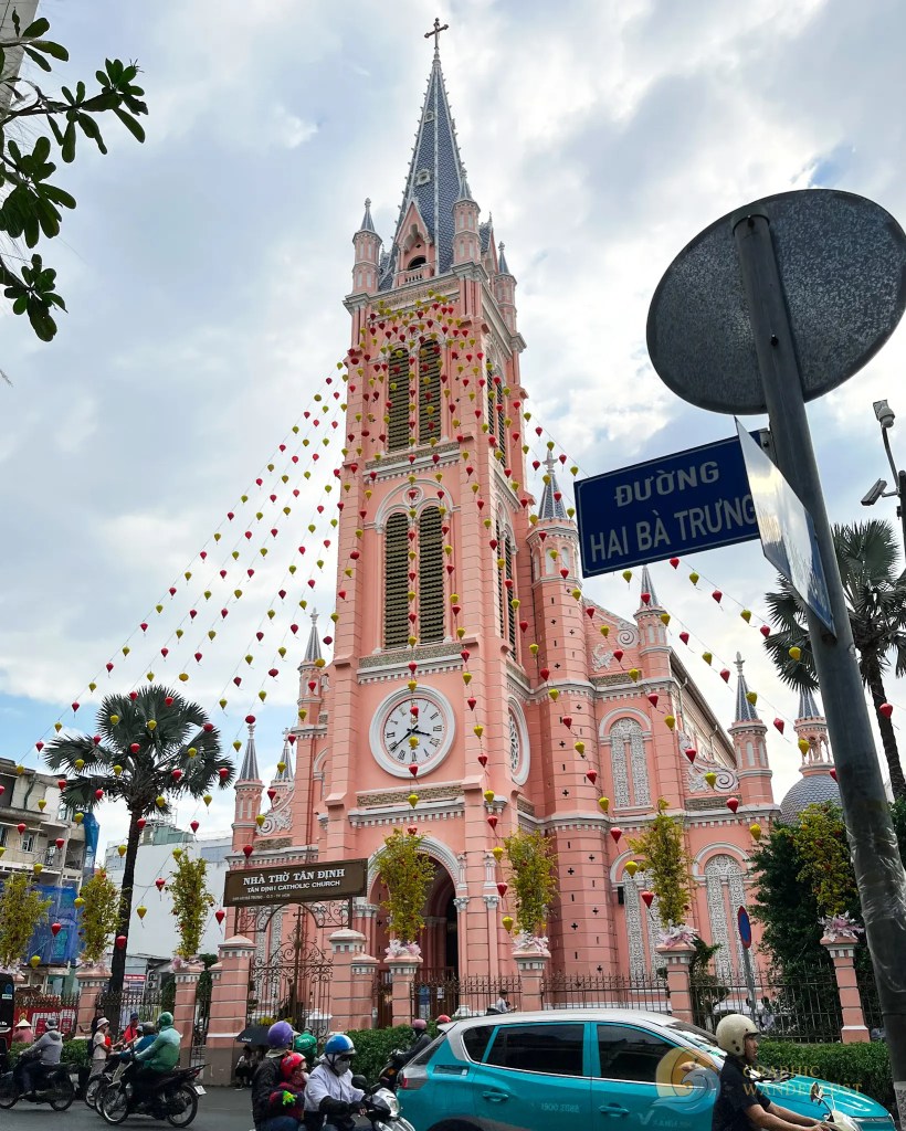 A pink church with a tall steeple adorned with colorful decorations, surrounded by palm trees and traffic, seen under a cloudy sky.