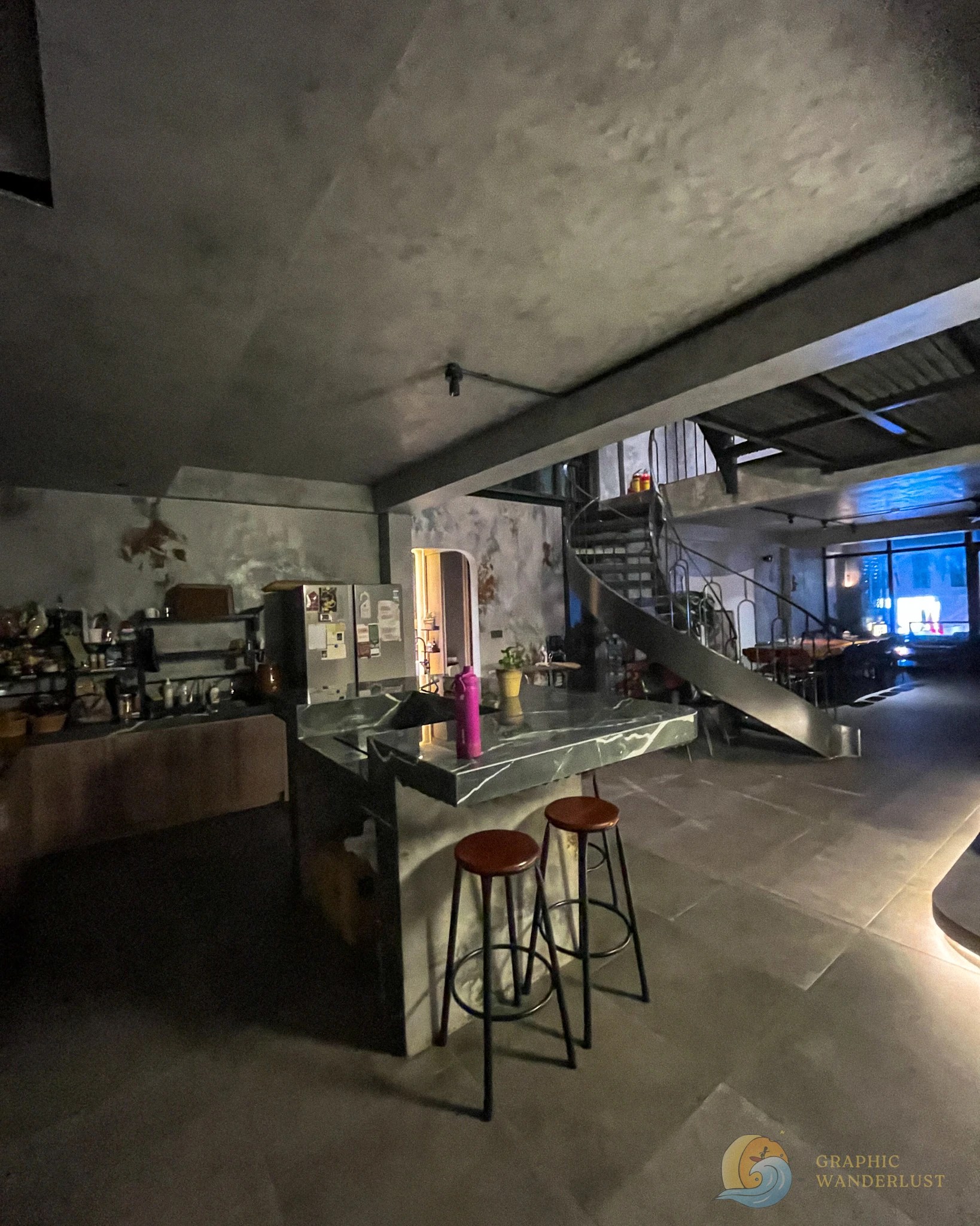 Interior of hostel boasting an industrial design with a kitchen island in the center and a dining table set at the farther end.
