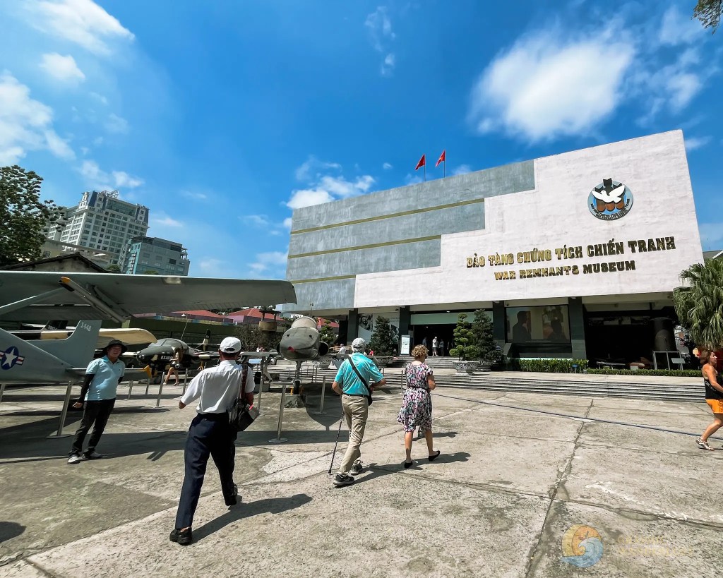 Visitors walking towards the War Remnants Museum, featuring military aircraft displayed outside against a clear blue sky.