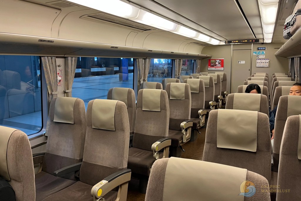 Interior of a passenger train car with beige seats and overhead luggage racks, two people seated on the right