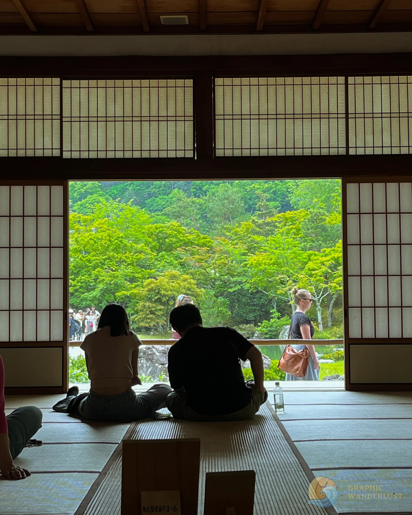 Two people sitting on a tatami mat inside a traditional Japanese building with a scenic view of lush greenery outside.