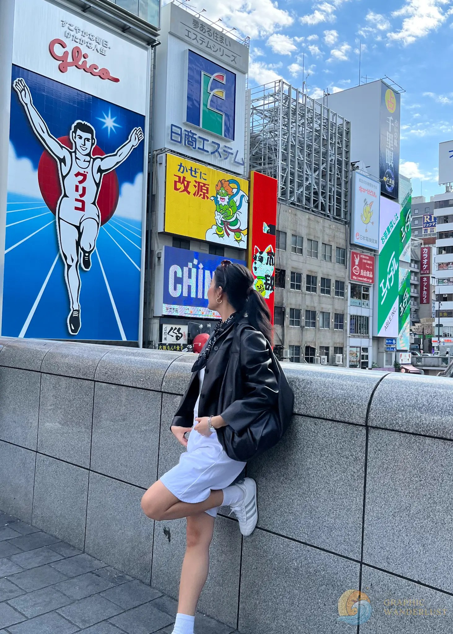 Woman leaning against stone railing looking at large colorful advertising signs on buildings in a cityscape under a partly cloudy sky