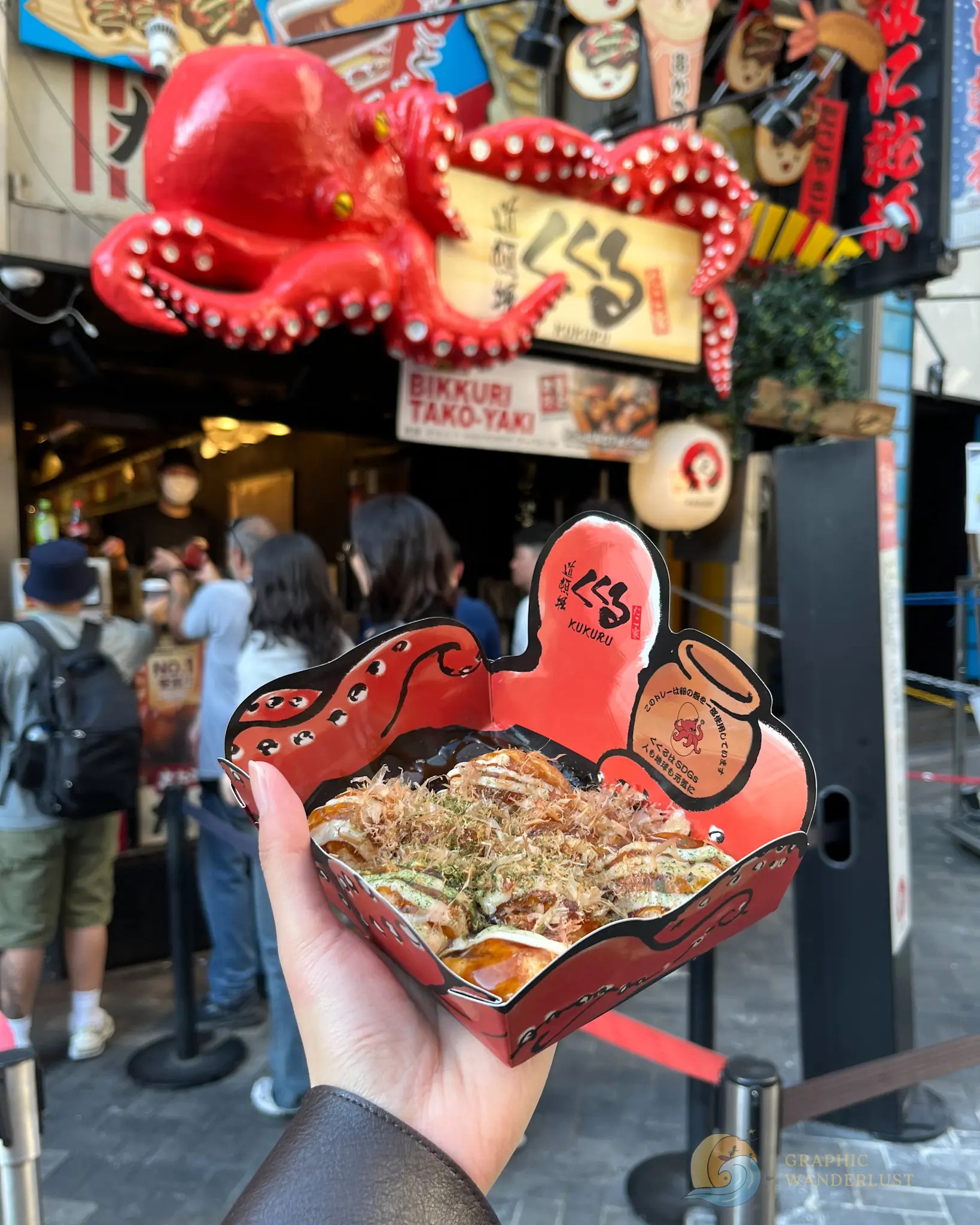 Kukuru Takoyaki Hand holding a container of takoyaki in front of a restaurant with a large red octopus sign and customers waiting