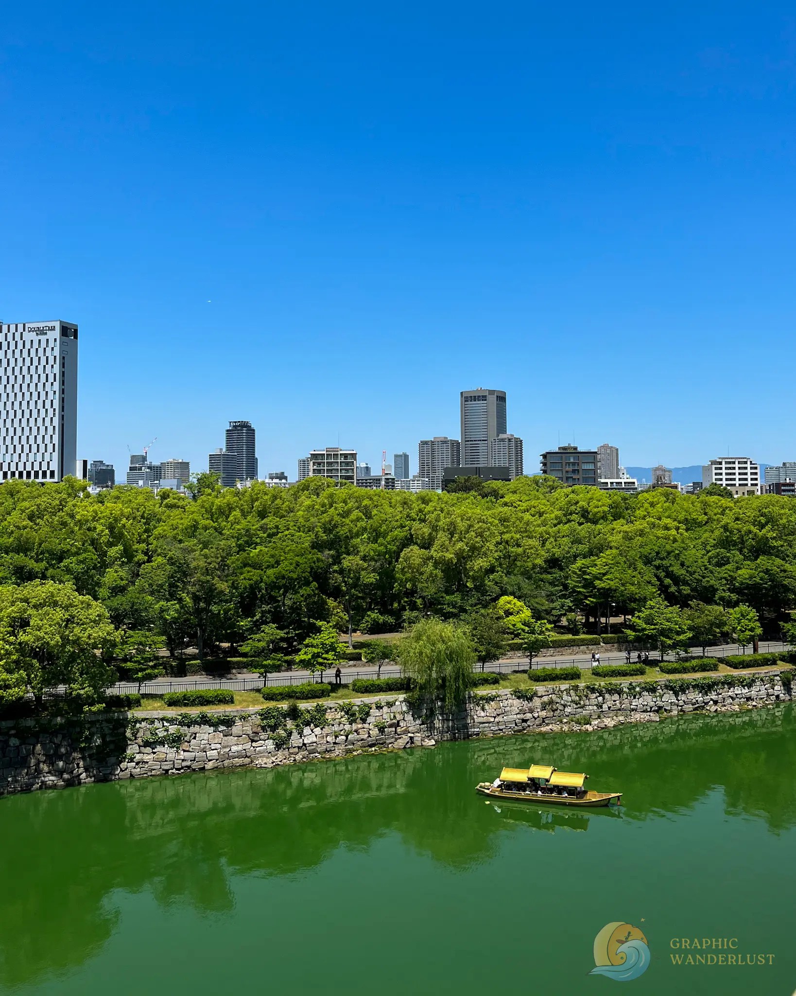 Osaka Castle Gozabune Boat A traditional wooden boat with passengers floats on a green river next to a stone wall and a green forest with a city skyline under a clear blue sky