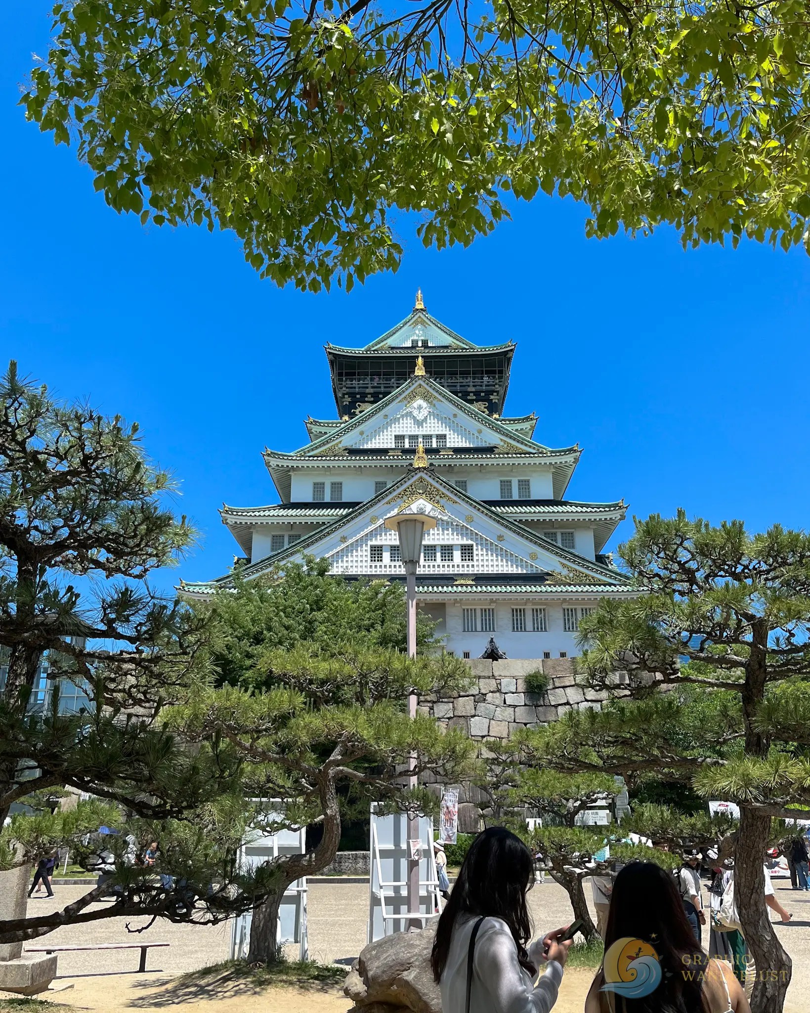 Osaka Castle Tower Front view of Osaka Castle with green trees and people in the foreground under a clear blue sky