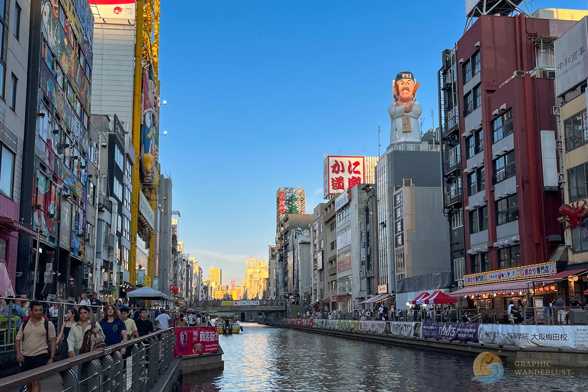 Tonbori River Walk Busy canal street with crowds, buildings, and colorful signs including a large Daruma doll and giant chef statue