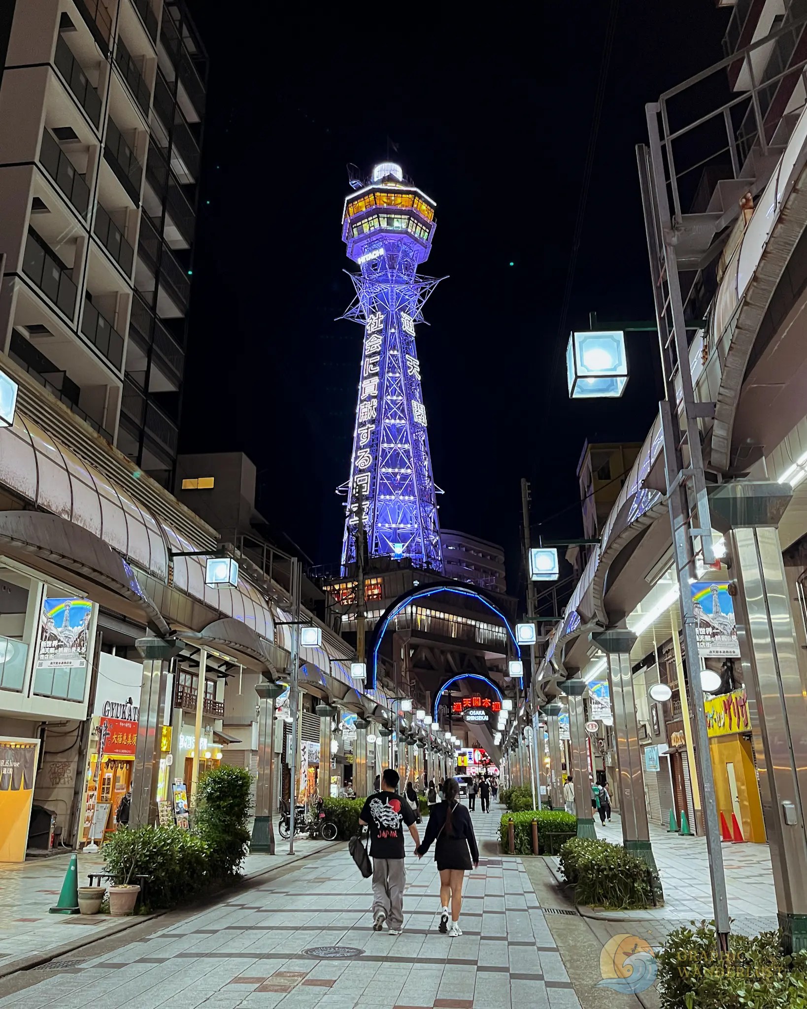 Tsutenkaku Tower Couple walking hand in hand along illuminated pedestrian street with shops and street lamps at night