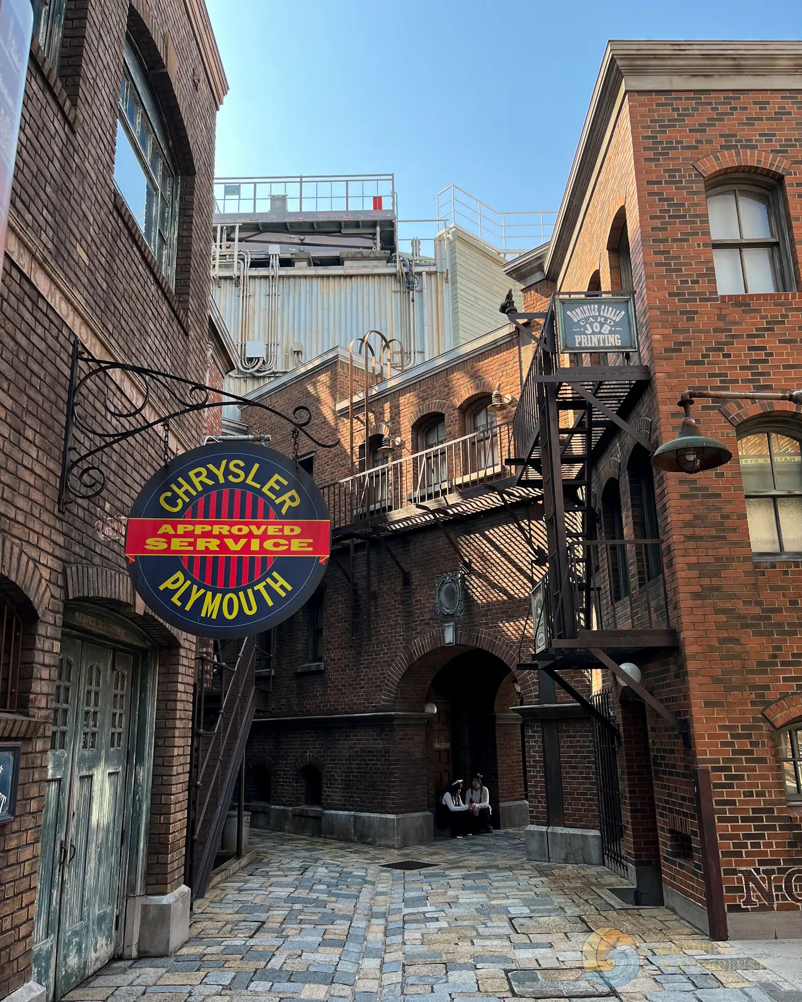 Narrow cobblestone alleyway with brick buildings, fire escapes, and vintage signs for Chrysler Plymouth service and card job printing