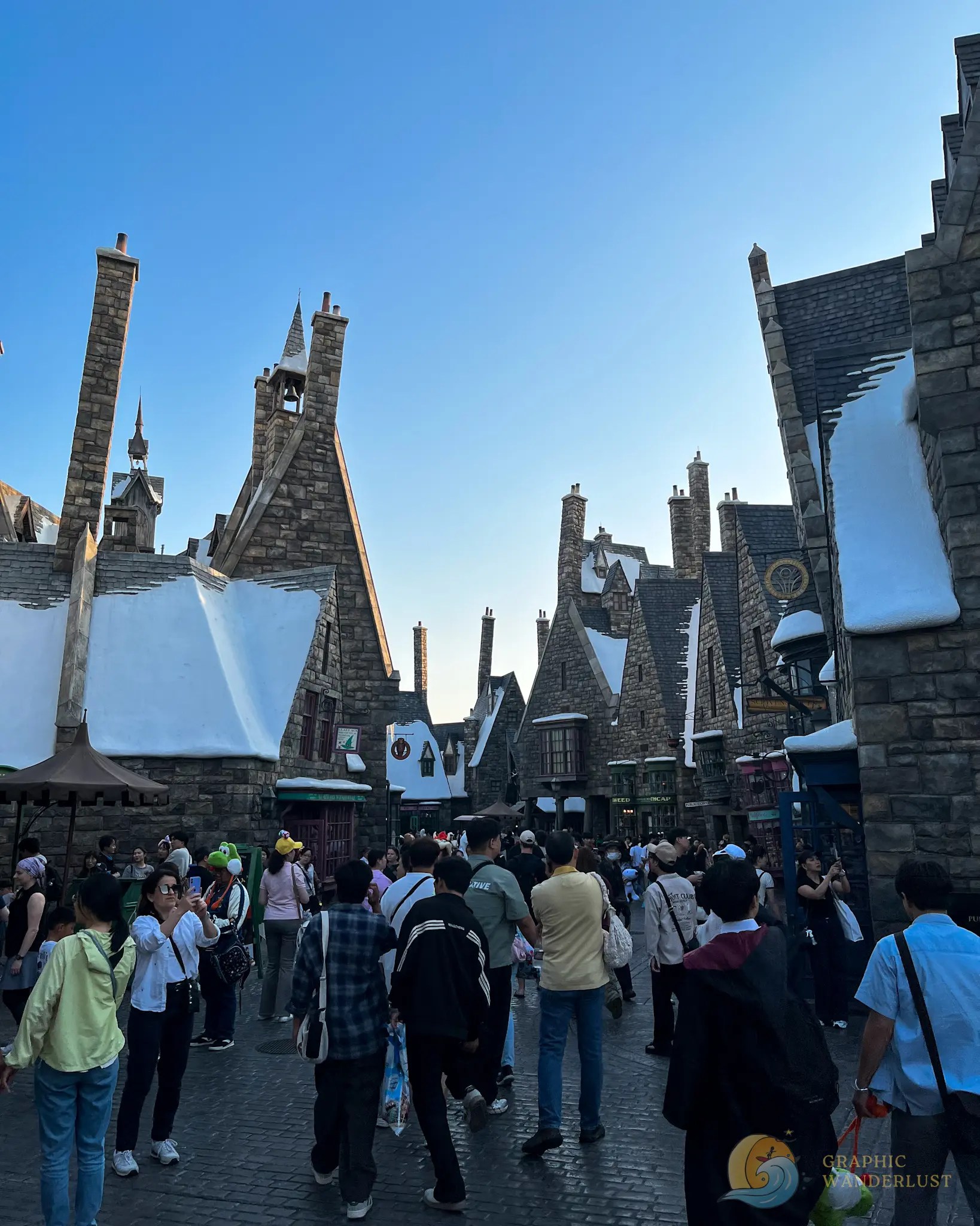 Crowd walking through a themed village street with snow-covered rooftops and stone chimneys under a clear blue sky