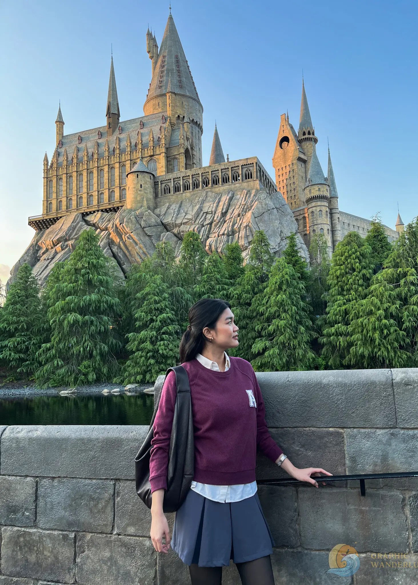 Woman standing near stone railing with Hogwarts castle and greenery in the background