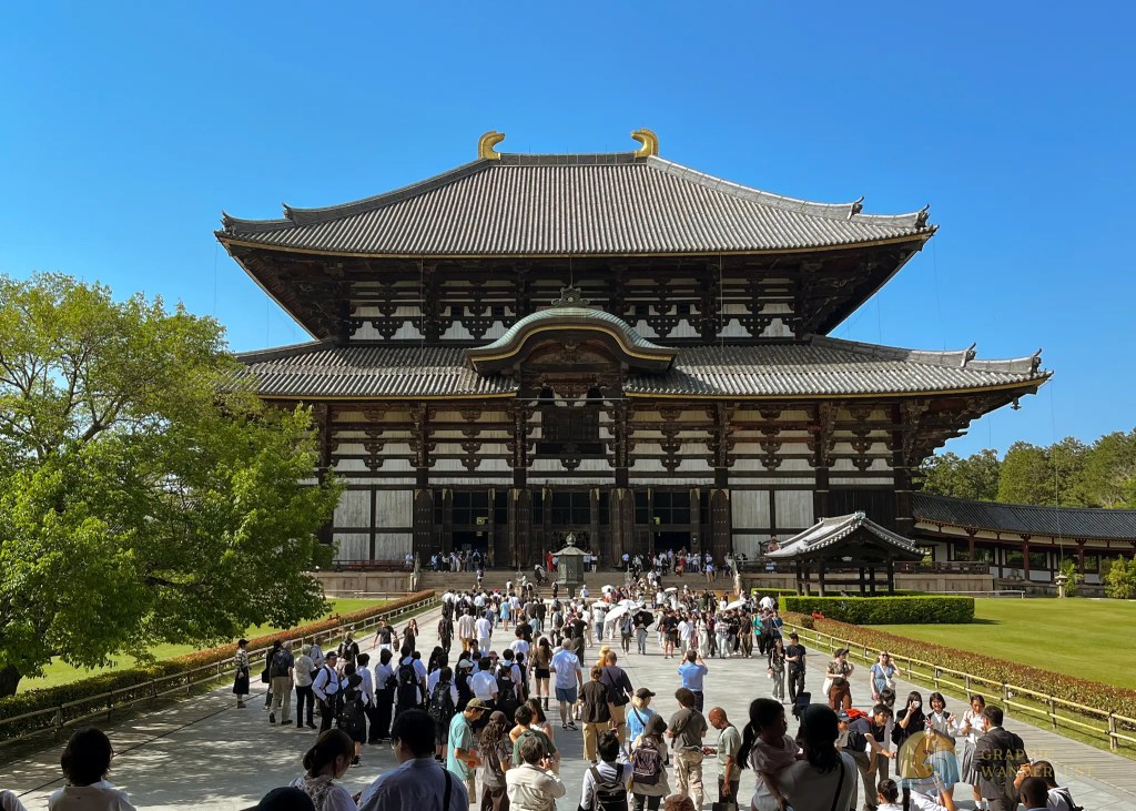 A big Buddhist temple with a clear blue sky in the background and a crowd of tourists walking towards it in its front.
