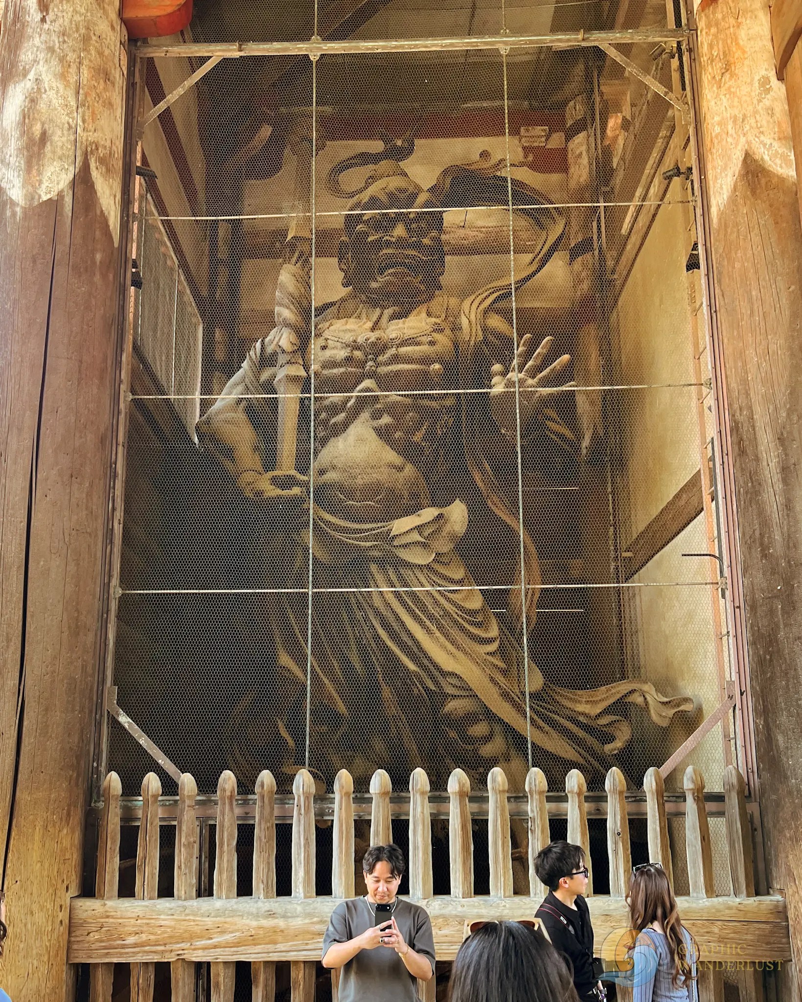 Large wooden statue with an angry expression behind protective wire mesh and wooden fence, with people standing in front