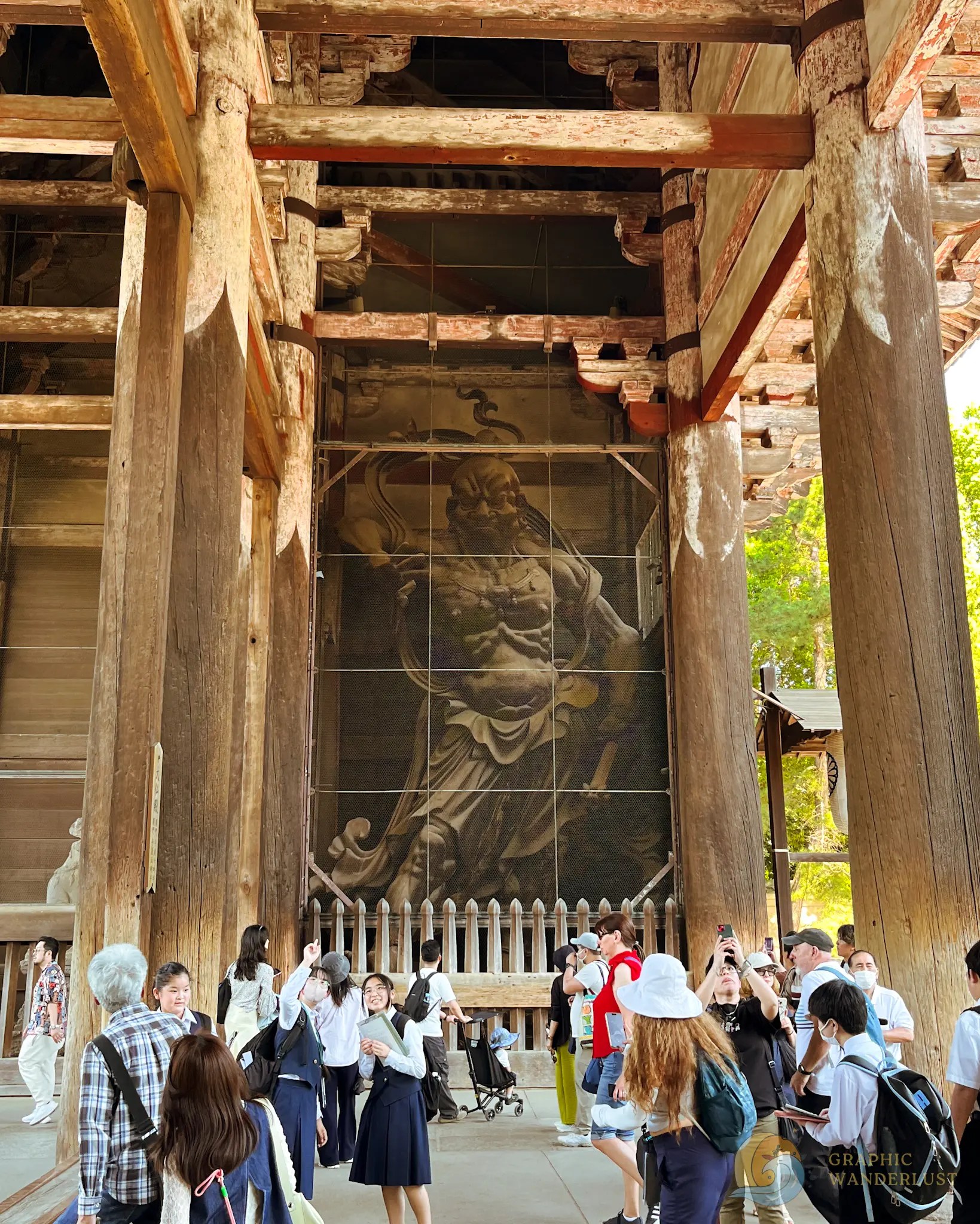 Tourists and students gathered inside a large wooden temple building with tall pillars and a giant statue behind a protective fence