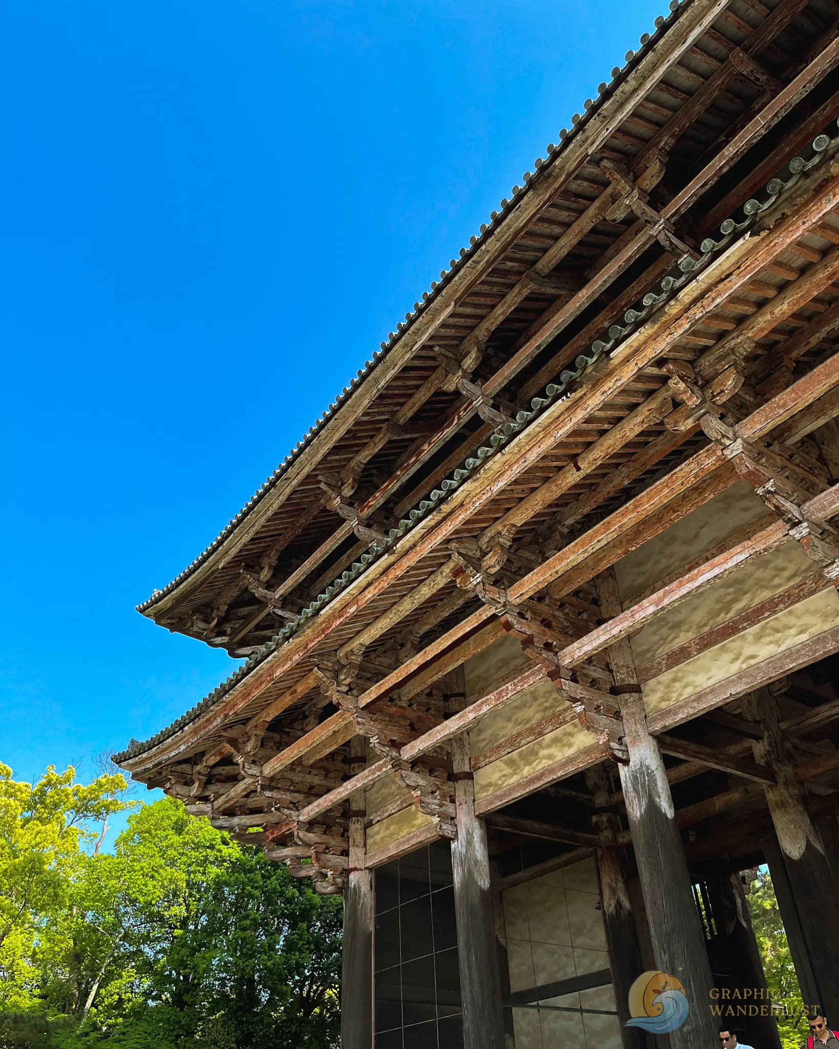 Wooden beams and roof structure of a traditional Japanese building with green trees and clear blue sky